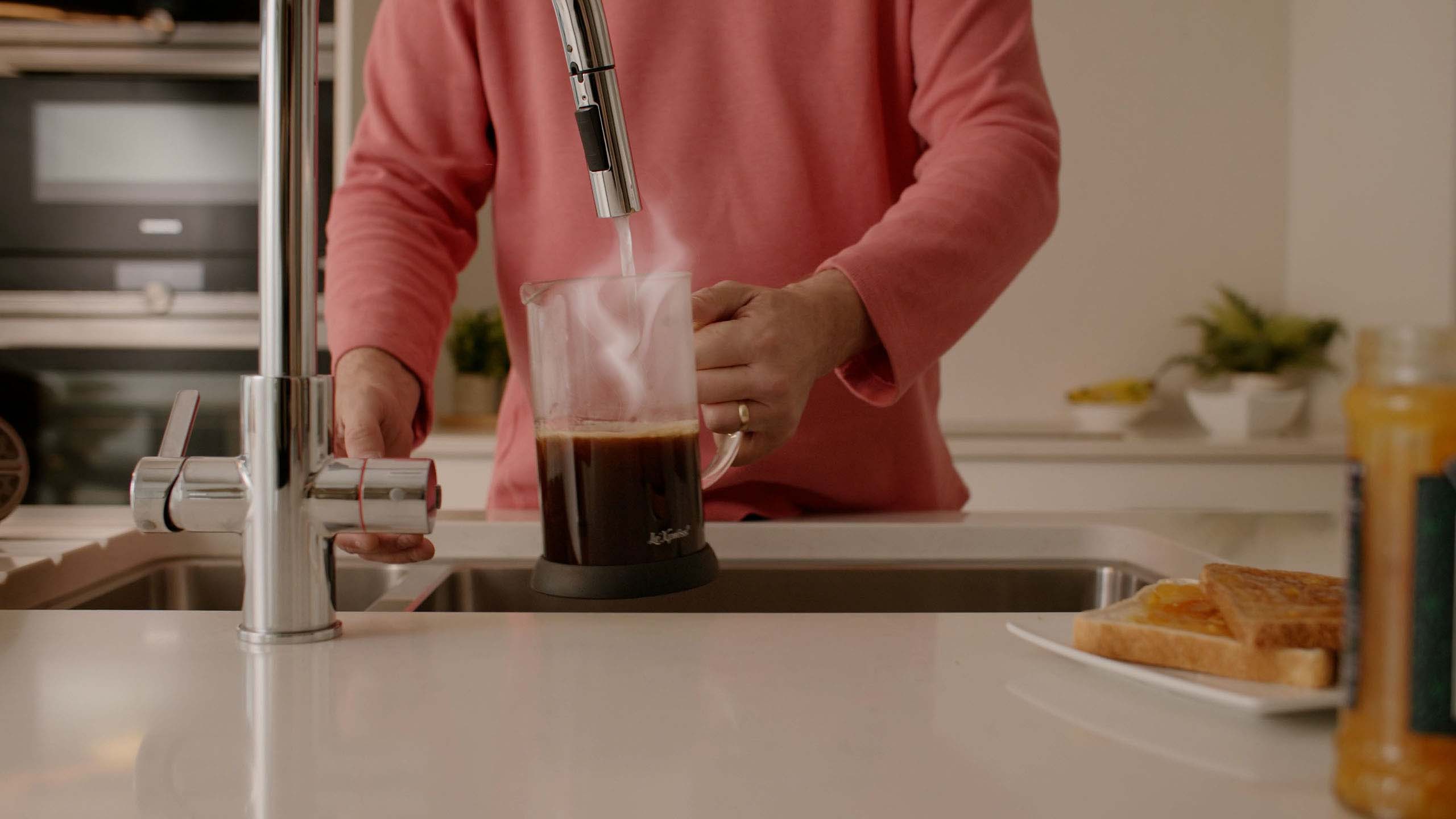 Person in red sweater filling a glass coffee press with hot water from a chrome kitchen tap