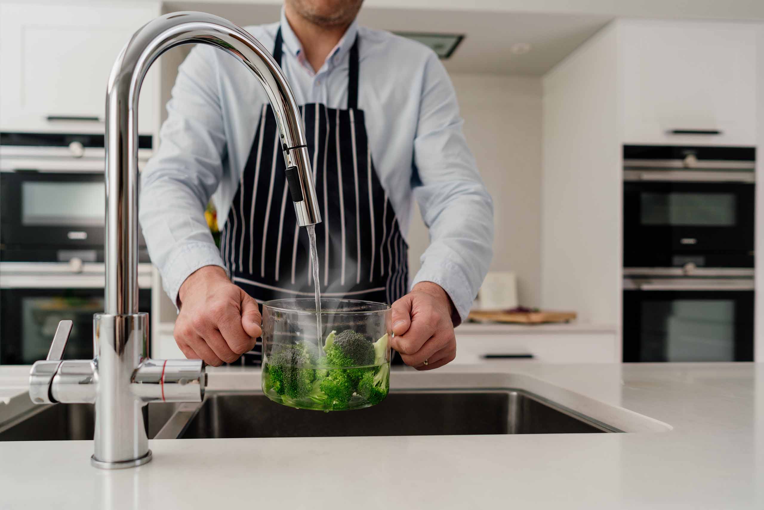 Man filling glass pot with broccoli under curved chrome kitchen tap over sink