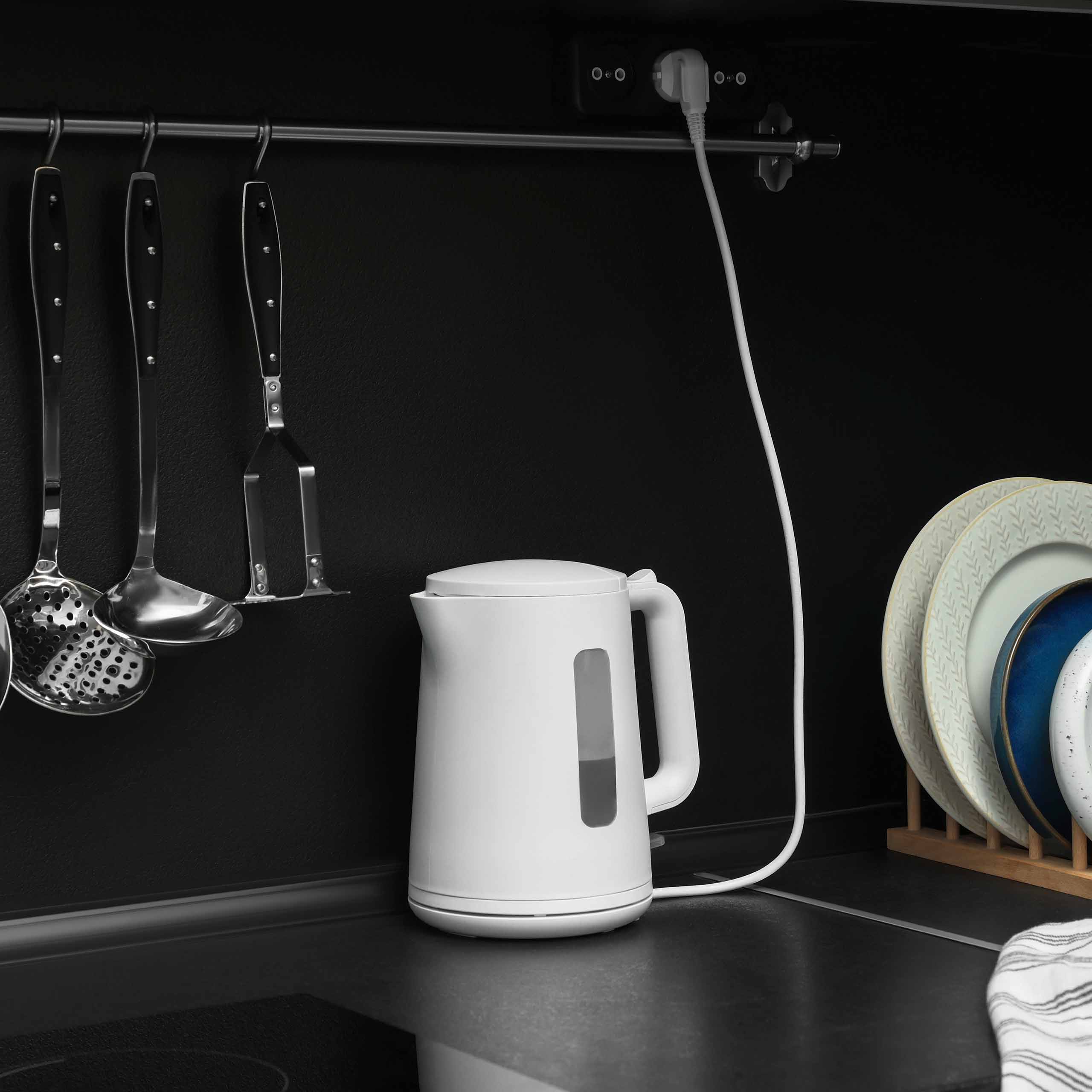 White electric kettle on black kitchen countertop next to hanging utensils and plates