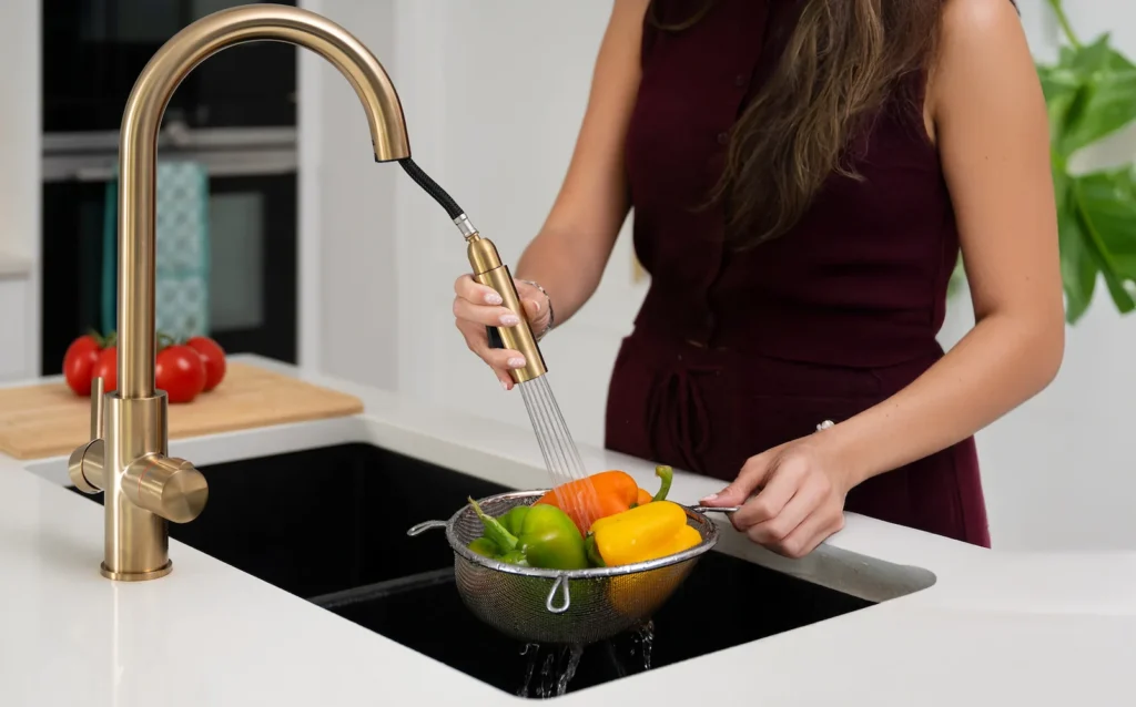 Brushed gold kitchen faucet with pull-down sprayer rinsing bell peppers in a metal strainer.