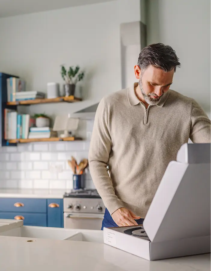 Person in beige sweater opening white box in a modern kitchen with blue cabinets.