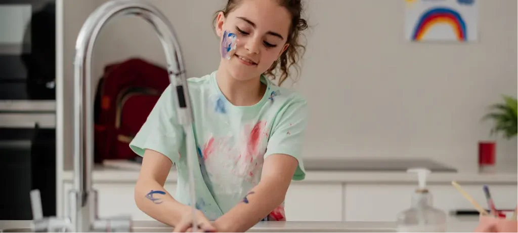 Child with paint on face and hands washing hands at kitchen sink