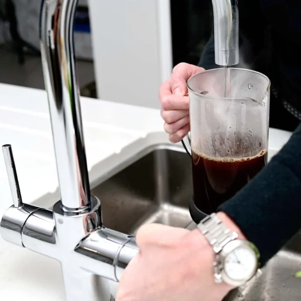 Quel Classic 4-in-1 instant boiling water tap in polished chrome, with a person dispensing boiling water into a glass coffee beaker over a stainless steel sink on a white worktop