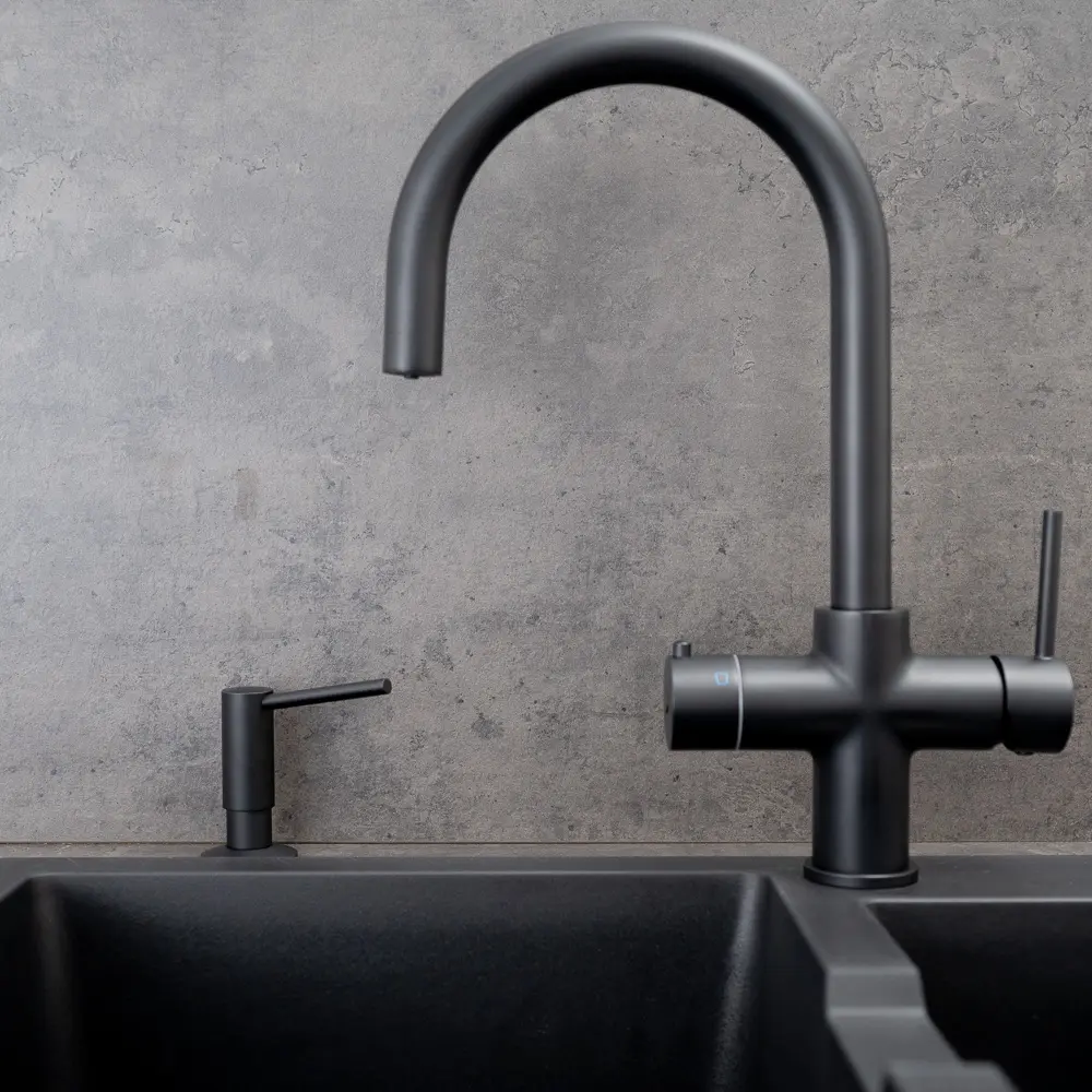 Quel Tap integrated soap dispenser in matt black, shown mounted beside a matching matt black Aquataps kitchen tap with a C-shaped spout and dual lever controls, both above a black granite double undermount sink against a dark concrete-effect splashback