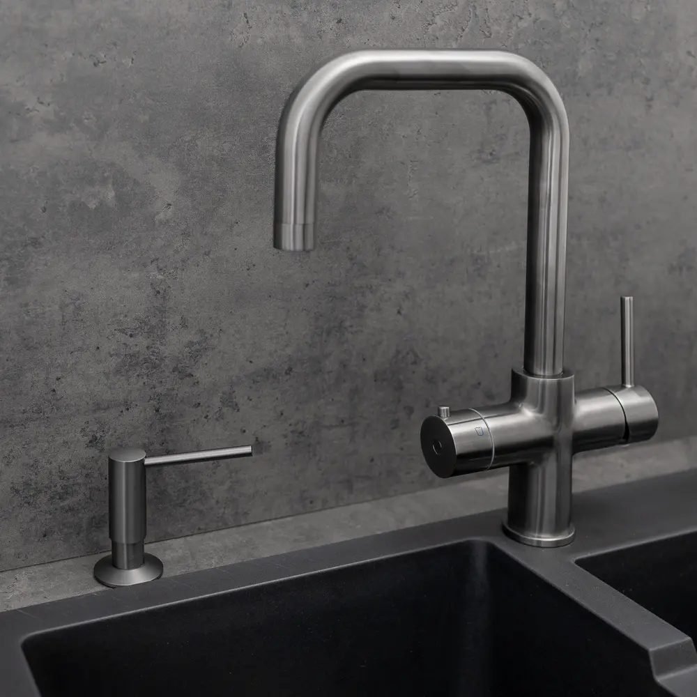 Quel Tap integrated soap dispenser in gunmetal grey, shown mounted beside a matching gunmetal grey Quel Tap kitchen tap with a square D-shaped spout, both above a black granite double undermount sink against a dark concrete-effect splashback