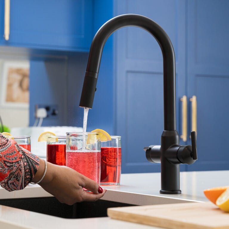 Quel Ultra Plus 4-in-1 instant boiling water tap in matt black, dispensing filtered water into a glass of red fruit drink with a lemon garnish in a modern blue kitchen.