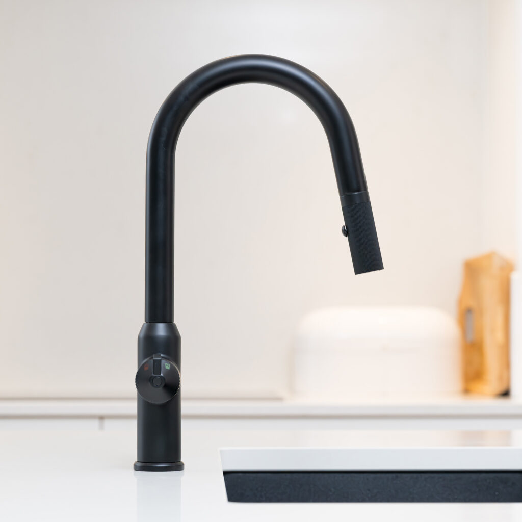 Quel Ultra Plus 4-in-1 instant boiling water tap in matt black, shown installed beside a black undermount sink with the pull-out spray head docked and multifunction control dial visible in a modern kitchen.