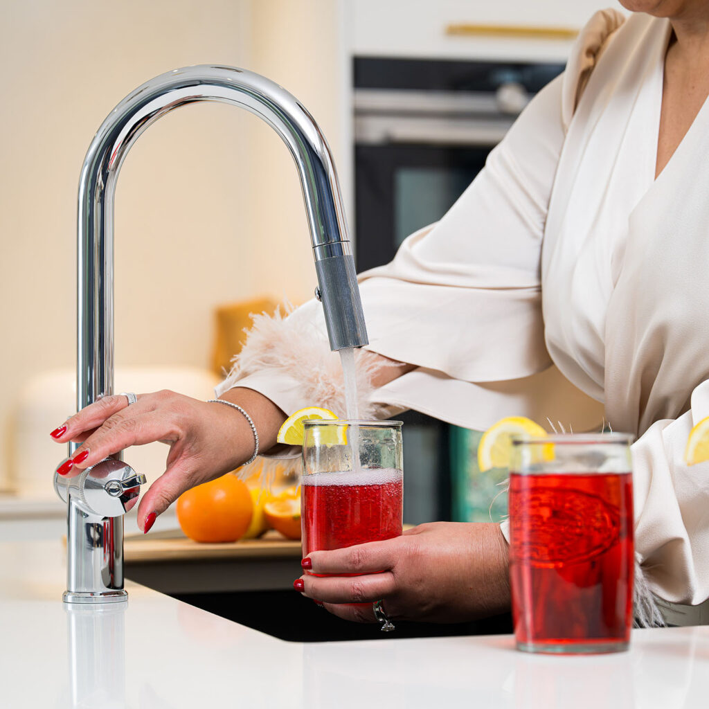 Quel Ultra Plus 4-in-1 instant boiling water tap in polished chrome, dispensing filtered water into a glass of red fruit drink with a lemon garnish, with the pull-out spray head docked in a modern kitchen.