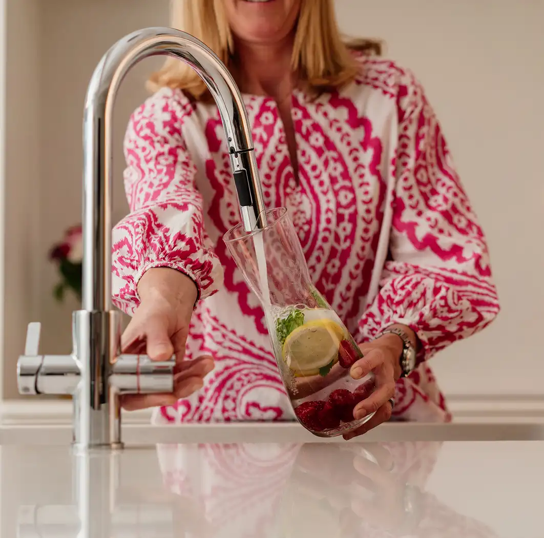 Person in patterned pink and white shirt filling glass with water from silver kitchen faucet