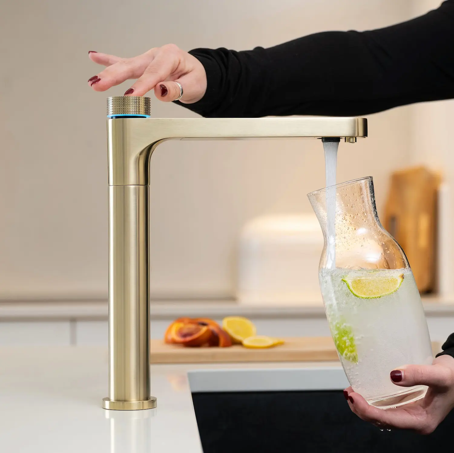 Quel Ultimo 6-in-1 instant boiling, chilled, sparkling and filtered water tap in brushed brass, dispensing sparkling water into a glass carafe with lime slices, as a person selects a mode using the knurled top knob with blue LED ring, in a warm neutral kitchen setting