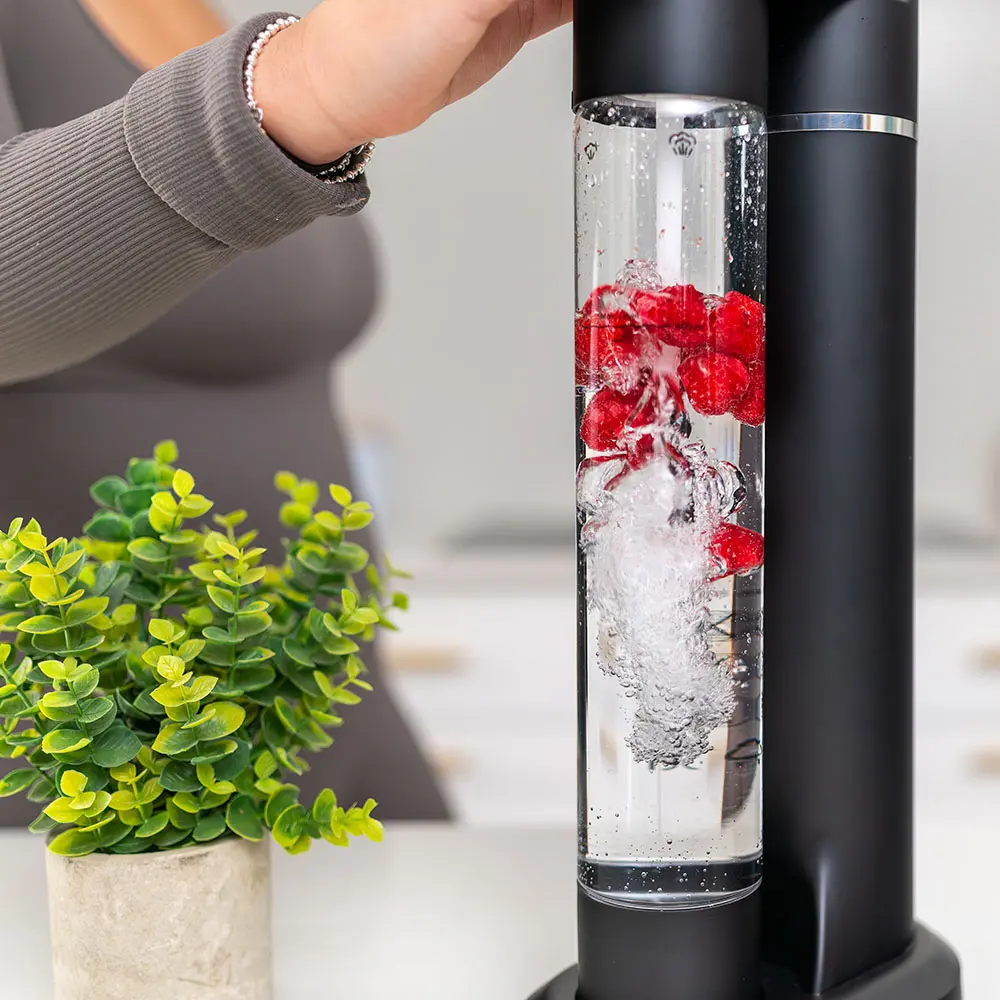 Close-up of the FizzyAqua sparkling water maker in matt black, showing the clear bottle actively carbonating raspberry-infused water with bubbles swirling around the fresh raspberries, with a small potted plant beside it