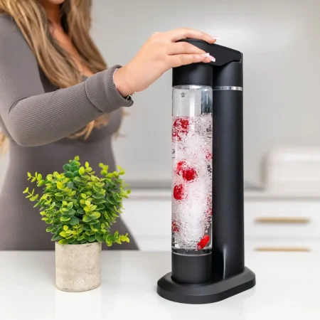 FizzyAqua sparkling water maker and fruit infuser in matt black, with a person pressing the carbonation button while the clear bottle is filled with sparkling water and infused with fresh raspberries, on a white worktop with a small potted plant beside it