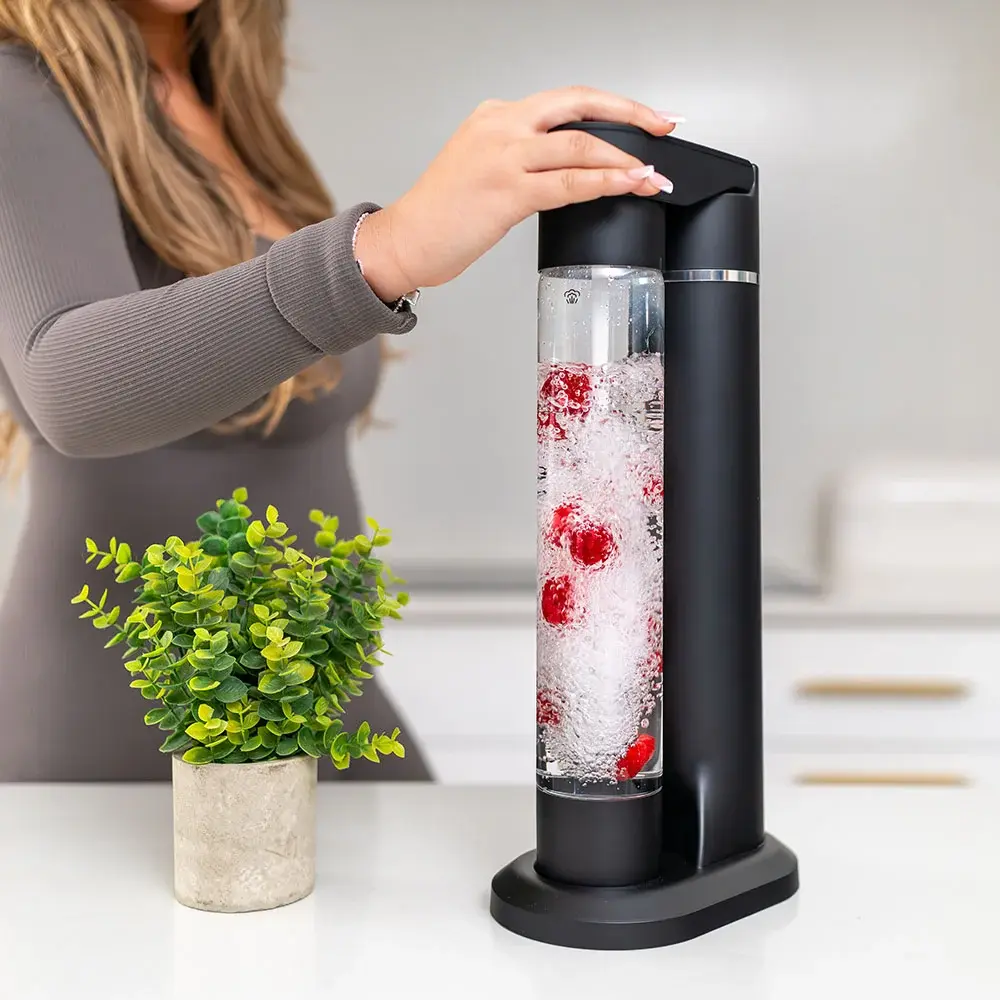 FizzyAqua sparkling water maker and fruit infuser in matt black, with a person pressing the carbonation button while the clear bottle is filled with sparkling water and infused with fresh raspberries, on a white worktop with a small potted plant beside it