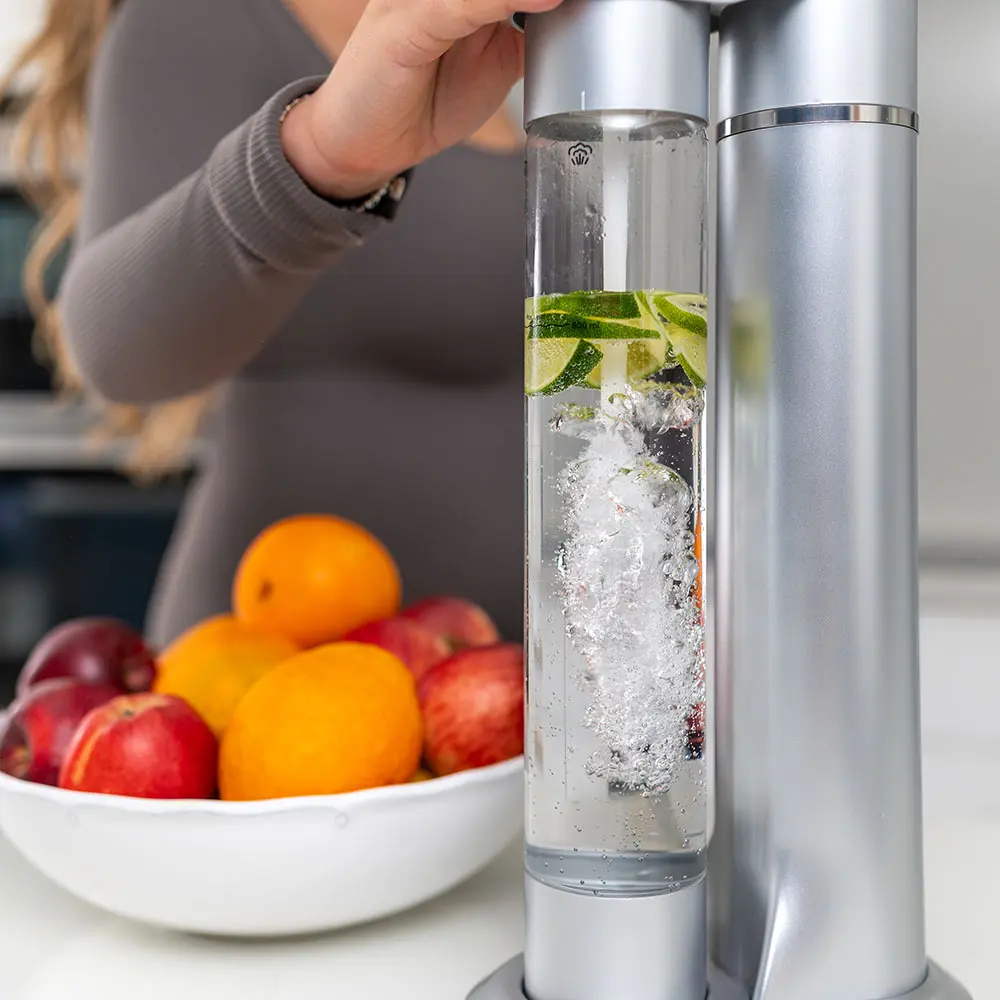 Close-up of the FizzyAqua sparkling water maker in silver, showing the clear bottle actively carbonating fruit-infused water with lime slices and berries, with a bowl of fresh oranges and apples visible in the background