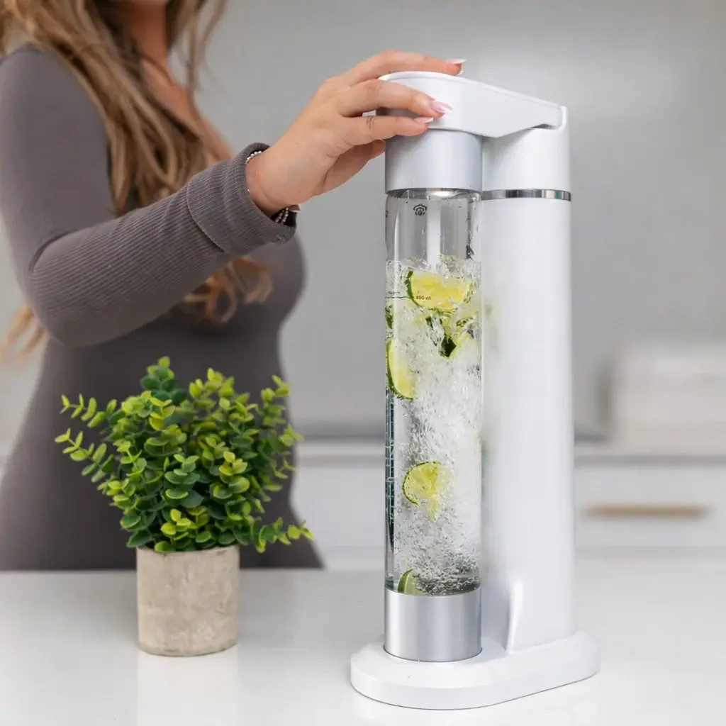 FizzyAqua sparkling water maker and fruit infuser in white, with a person pressing the carbonation button while the clear bottle is filled with sparkling water, ice and lime slices, on a white worktop with a small potted plant in the background