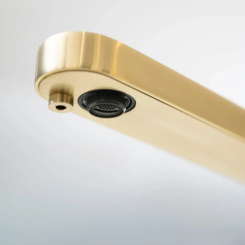 Underside close-up of the Quel Ultimo tap spout in brushed brass, showing the dual outlets — a mesh aerator and a separate smaller nozzle — set against a plain white background
