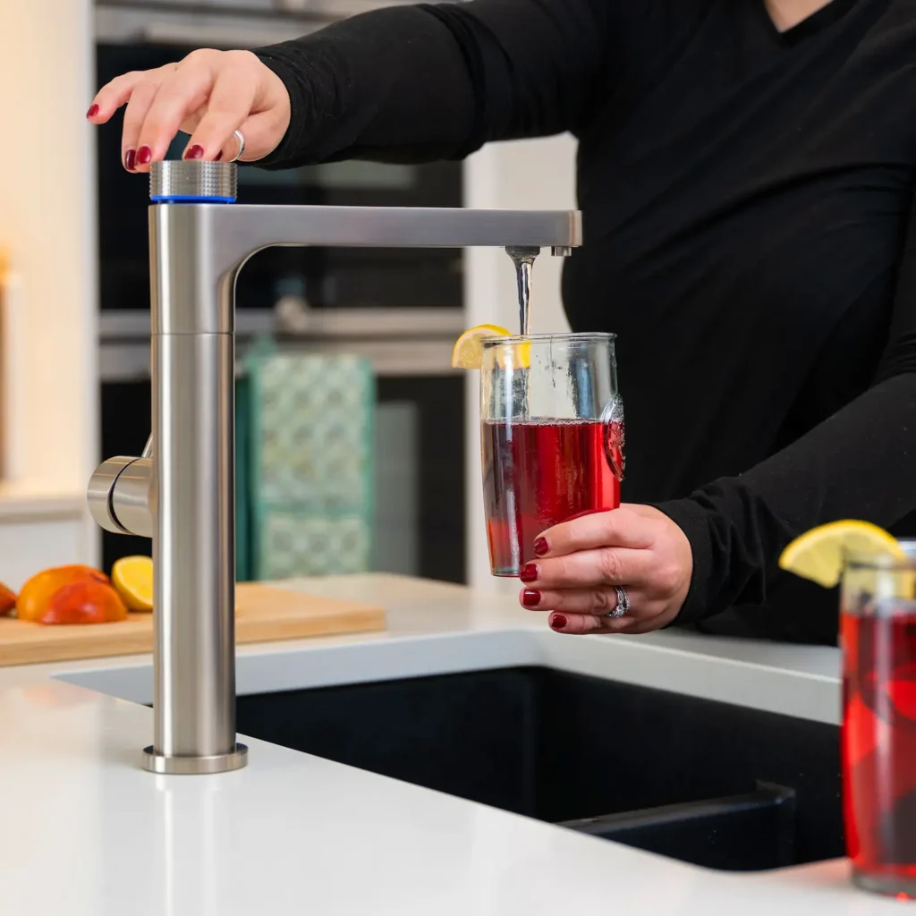 Quel Ultimo 6-in-1 instant boiling, chilled, sparkling and filtered water tap in brushed steel, dispensing chilled filtered water into a glass of red fruit drink with a lemon slice, as a person selects a mode using the knurled top knob with blue LED ring, with fresh fruit and a second drink visible in the background