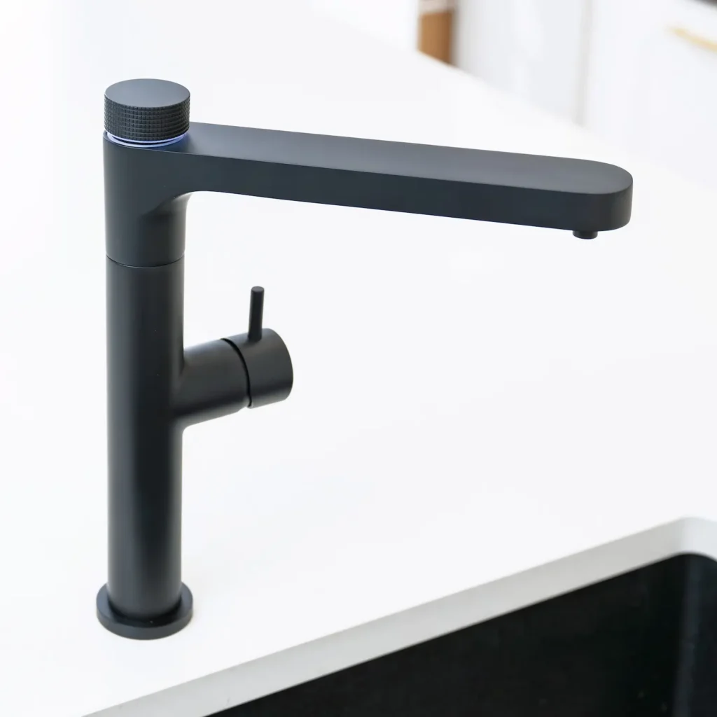 Quel Ultimo 6-in-1 instant boiling, chilled, sparkling and filtered water tap in matt black, shown from a side angle on a white kitchen worktop beside a black undermount sink, against a clean white background