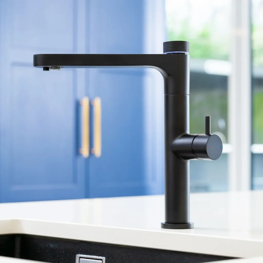 Quel Ultimo 6-in-1 instant boiling, chilled, sparkling and filtered water tap in matt black, mounted on a white kitchen worktop with a black sink, set against blue cabinetry with gold handles