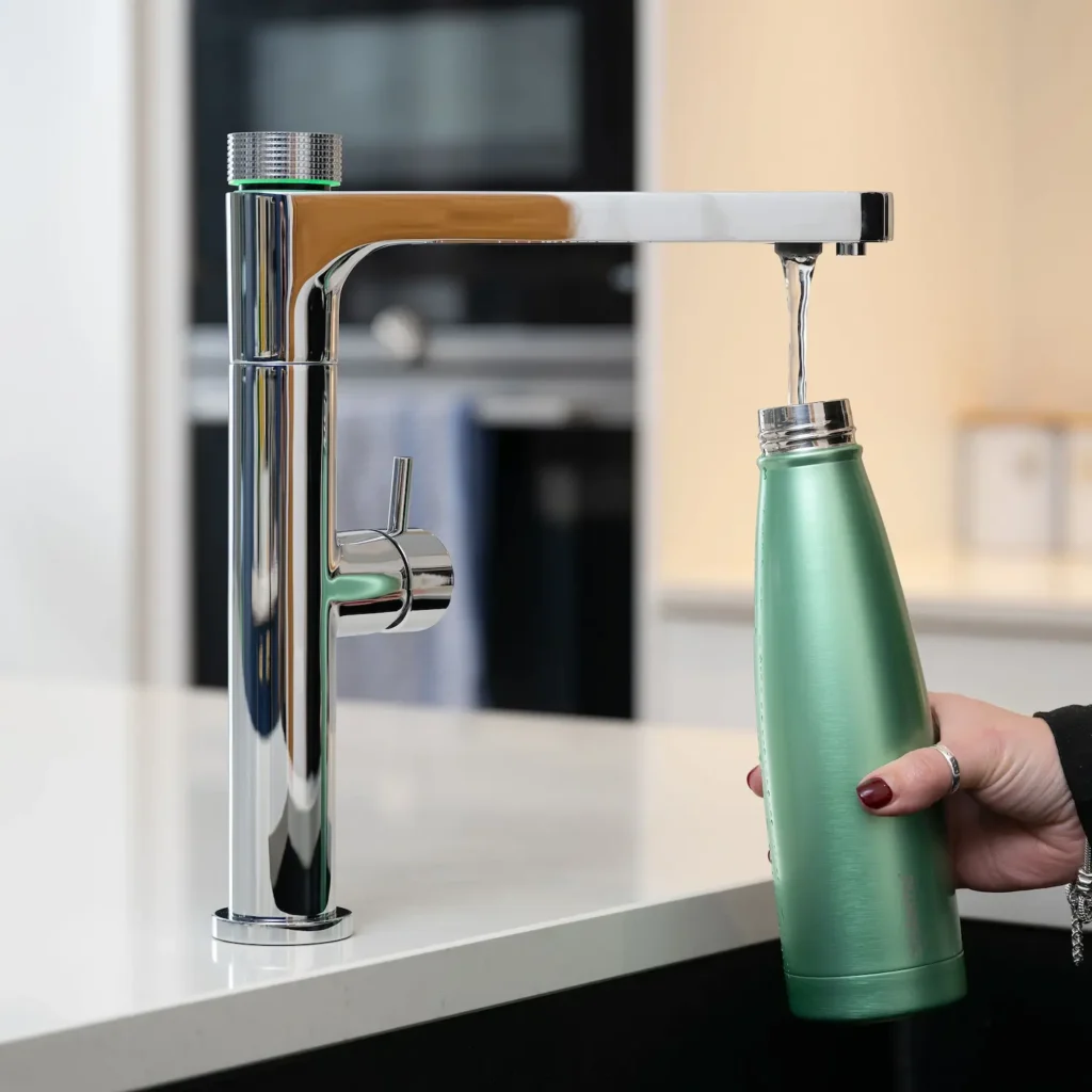 Quel Ultimo 6-in-1 instant boiling, chilled, sparkling and filtered water tap in chrome, dispensing filtered water into a green reusable bottle held by a person's hand, mounted on a white kitchen worktop with an oven visible in the background