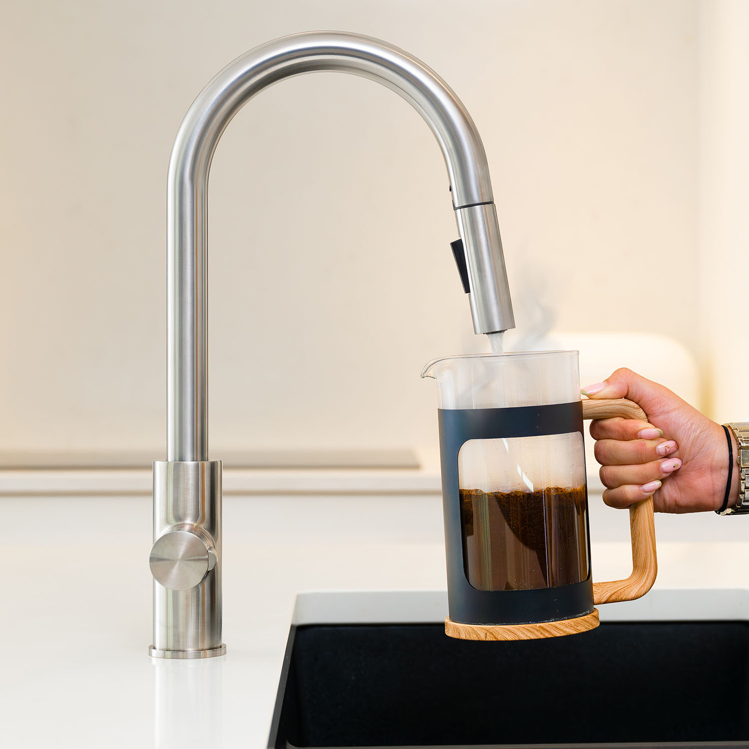 Quel Ultra 4-in-1 instant boiling water tap in brushed nickel, dispensing boiling water into a glass cafetière, with the pull-out spray head docked in a modern blue kitchen.