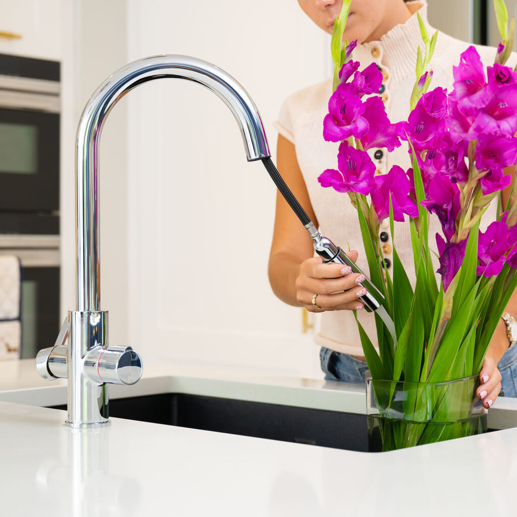 Quel Ultra 4-in-1 instant boiling water tap in polished chrome, shown using the extended pull-out spray head to fill a glass vase with pink gladiolus flowers beside a black undermount sink.