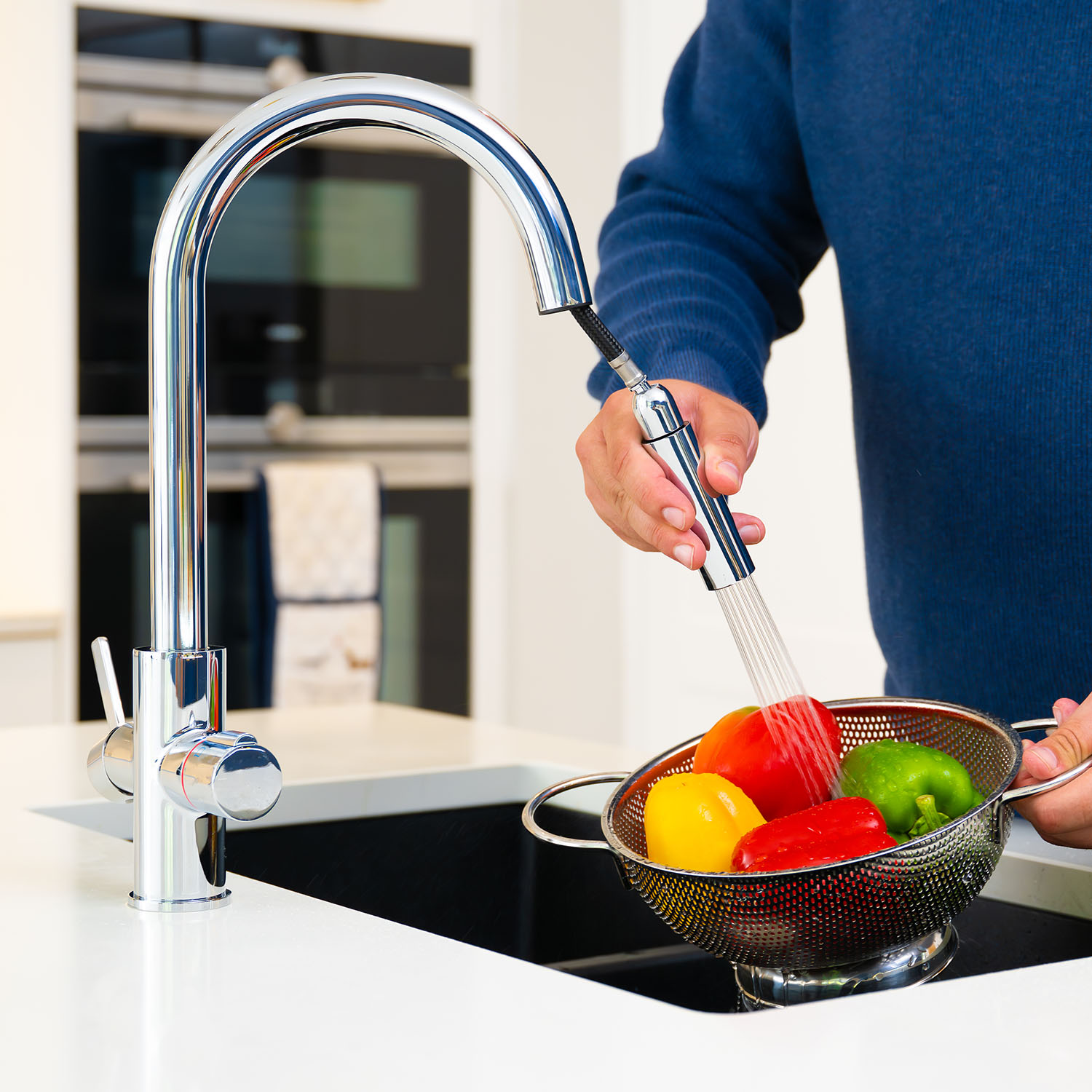 Quel Ultra 4-in-1 instant boiling water tap in polished chrome, shown using the extended pull-out spray head to rinse fresh peppers in a colander over a black undermount sink.