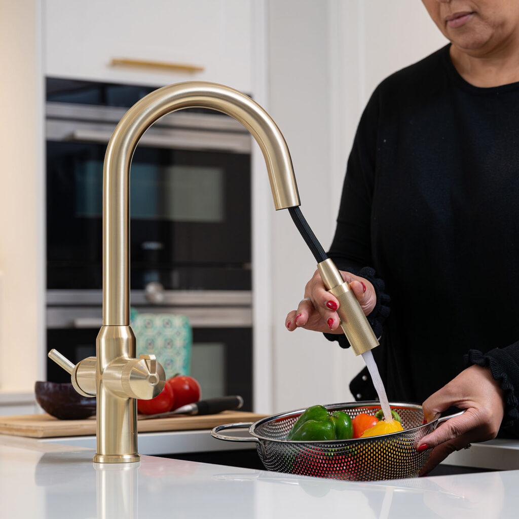 Quel Ultra Plus 4-in-1 instant boiling water tap in brushed brass, shown using the extended pull-out spray head to rinse fresh peppers in a colander over a black undermount sink.